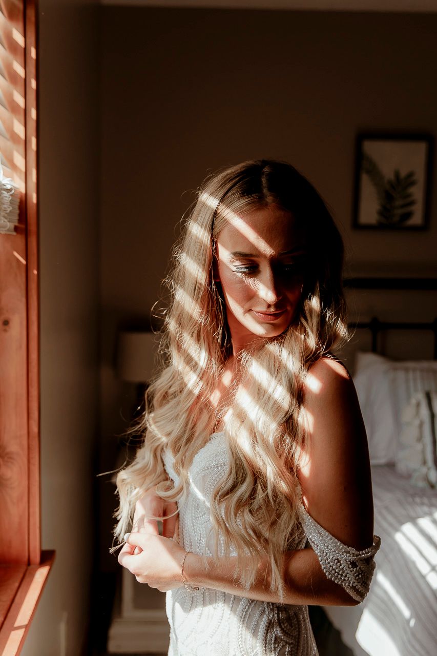 Sunlight through window blinds creates dramatic shadows on figure in white lace dress standing in bedroom.