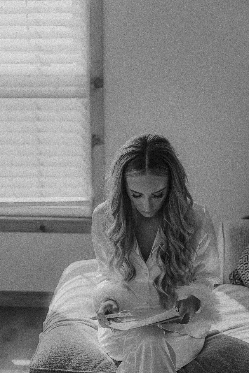 A person in a white robe reads while sitting on a bed near a window with natural light streaming in, captured in black and white.