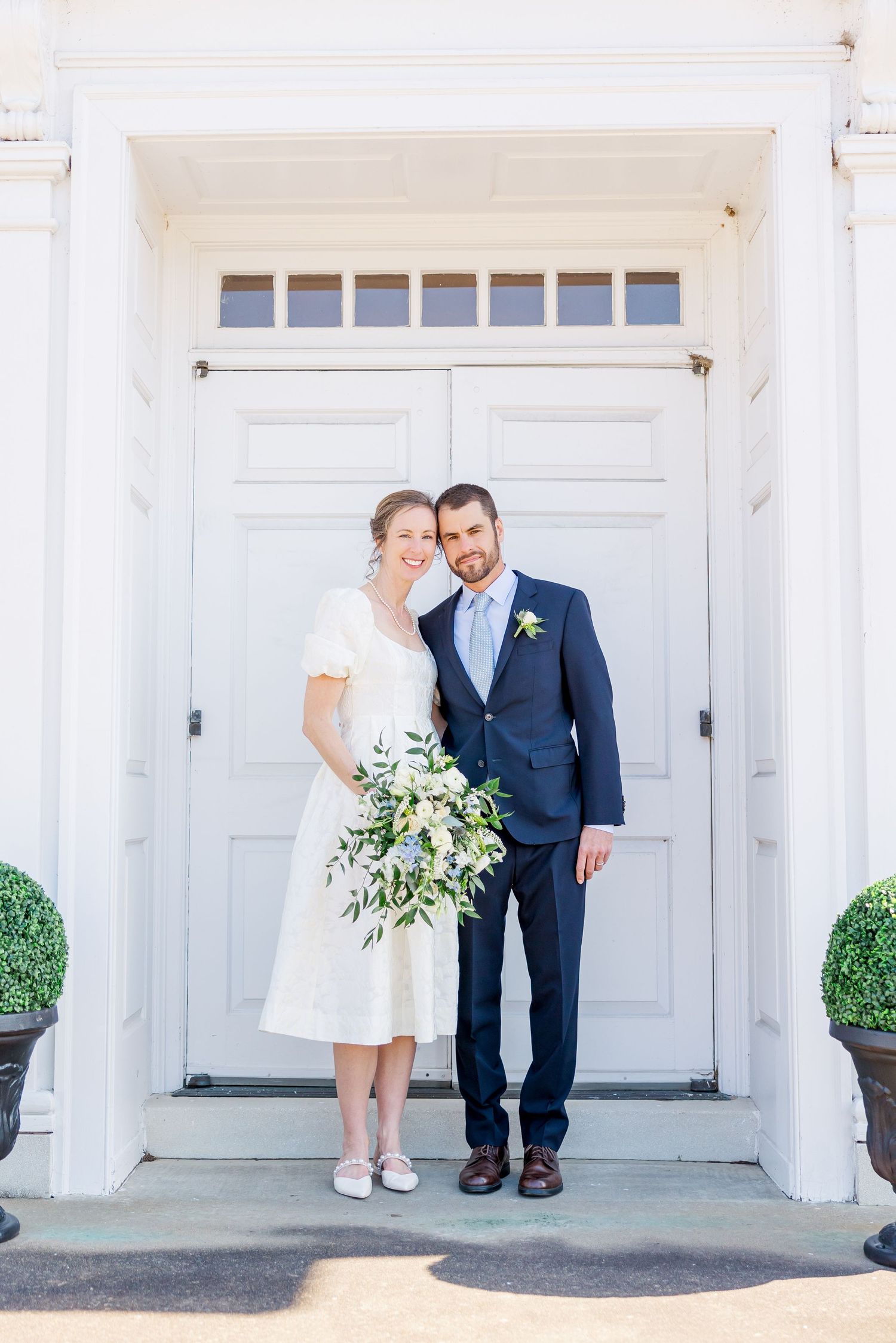 A couple poses in front of a grand white door with transom windows, bride holding a greenery bouquet.