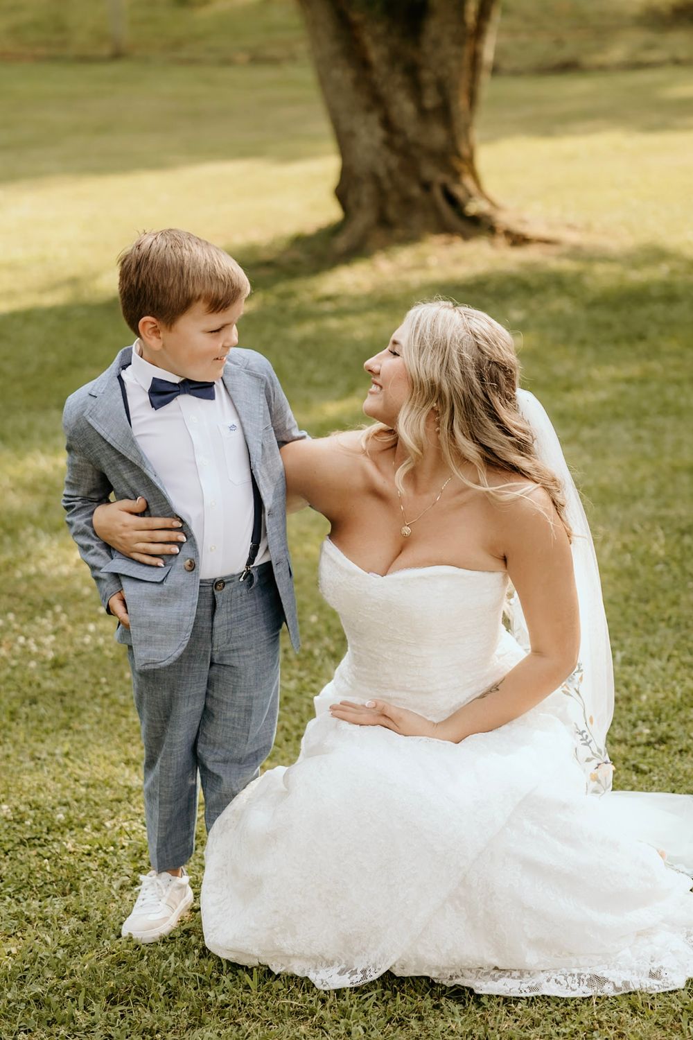 A touching wedding moment outdoors on grass with a bride in white dress and young boy wearing suspenders and bowtie.