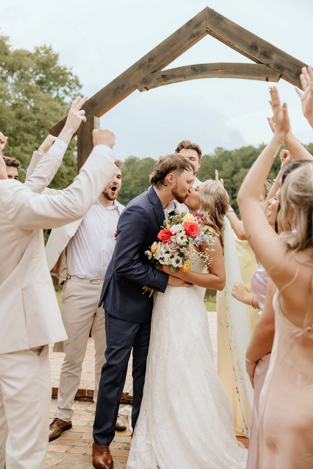 Couple sharing a romantic kiss at outdoor wedding ceremony while guests celebrate beneath rustic wooden arch.