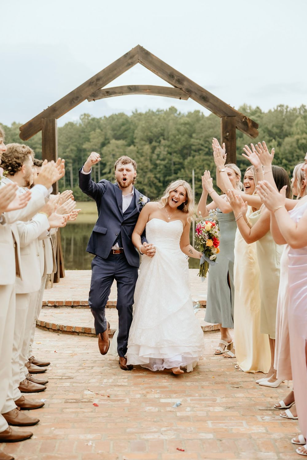 Newlyweds celebrate joyfully under wooden arch as wedding party in pastel dresses cheers them on during outdoor ceremony.