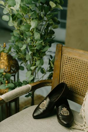 Vintage black leather shoes with ornate brooches displayed on a chair beside green eucalyptus plants and decorative elements.