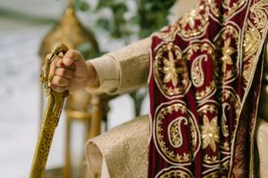Ornate gold staff held by hands wearing embroidered cream garments against deep red and gold religious vestments.