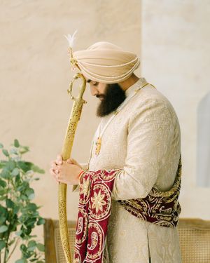 Traditional South Asian groom in cream sherwani and burgundy embroidered stole holds ornate gold staff in elegant portrait.