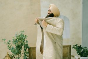 A person in a cream colored traditional sherwani adjusts their tie while standing near green plants outdoors.