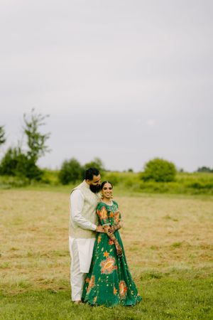 Couple in traditional Indian attire embracing in a green field under overcast sky.