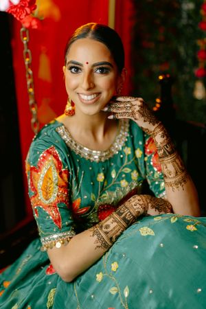 Traditional Indian bride wearing a teal and orange embroidered lehenga displays intricate henna designs on her hands.