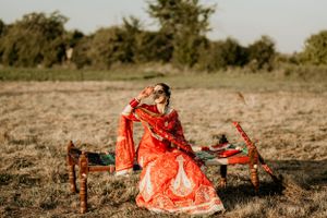 A traditional Indian bridal outfit in bright orange and gold lengha photographed outdoors in a rustic field setting.
