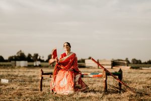 Person in a salmon-colored traditional Indian outfit sitting on a rustic wooden fence in an open field.