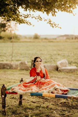 Traditional Indian attire in vibrant red and gold photographed during sunset with rustic outdoor setting and decorative furniture.