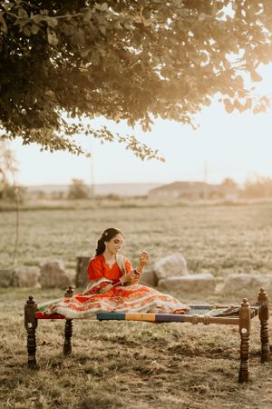 A traditional Indian charpai bed sits in a golden field at sunset with someone in an orange and red dress sitting upon it.