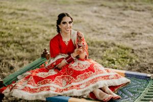 Bride in red and white lehenga poses outdoors on a blanket with traditional wedding accessories.