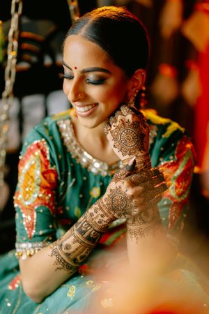 Bride in vibrant green and orange lehenga smiles while showing her detailed henna mehndi designs on hands.