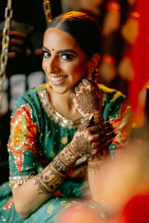 Indian bride in green and gold lehenga shows intricate henna designs on her hands during wedding celebrations.
