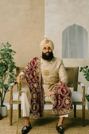 Traditional South Asian groom seated in formal attire wearing cream sherwani and burgundy embroidered shawl.