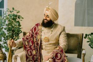 A person wearing a beige and gold traditional Indian wedding sherwani with an ornate burgundy shawl sits on a chair.