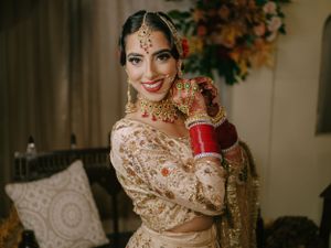 Traditional Indian bride in an ornate beige lehenga with intricate embroidery and jewelry poses elegantly indoors.