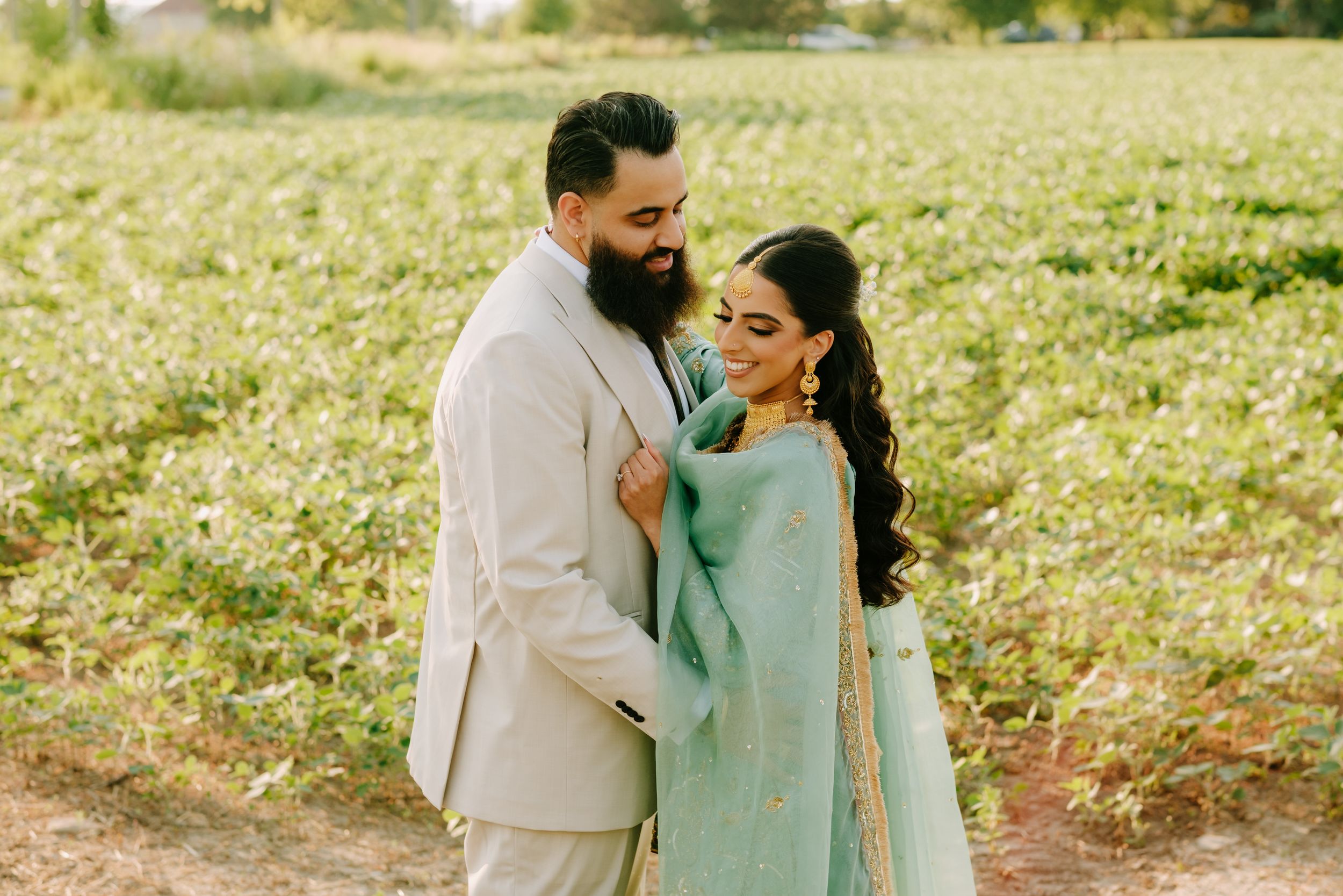A romantic couple embraces in a sunlit field wearing elegant ethnic attire during a sunny outdoor photoshoot.