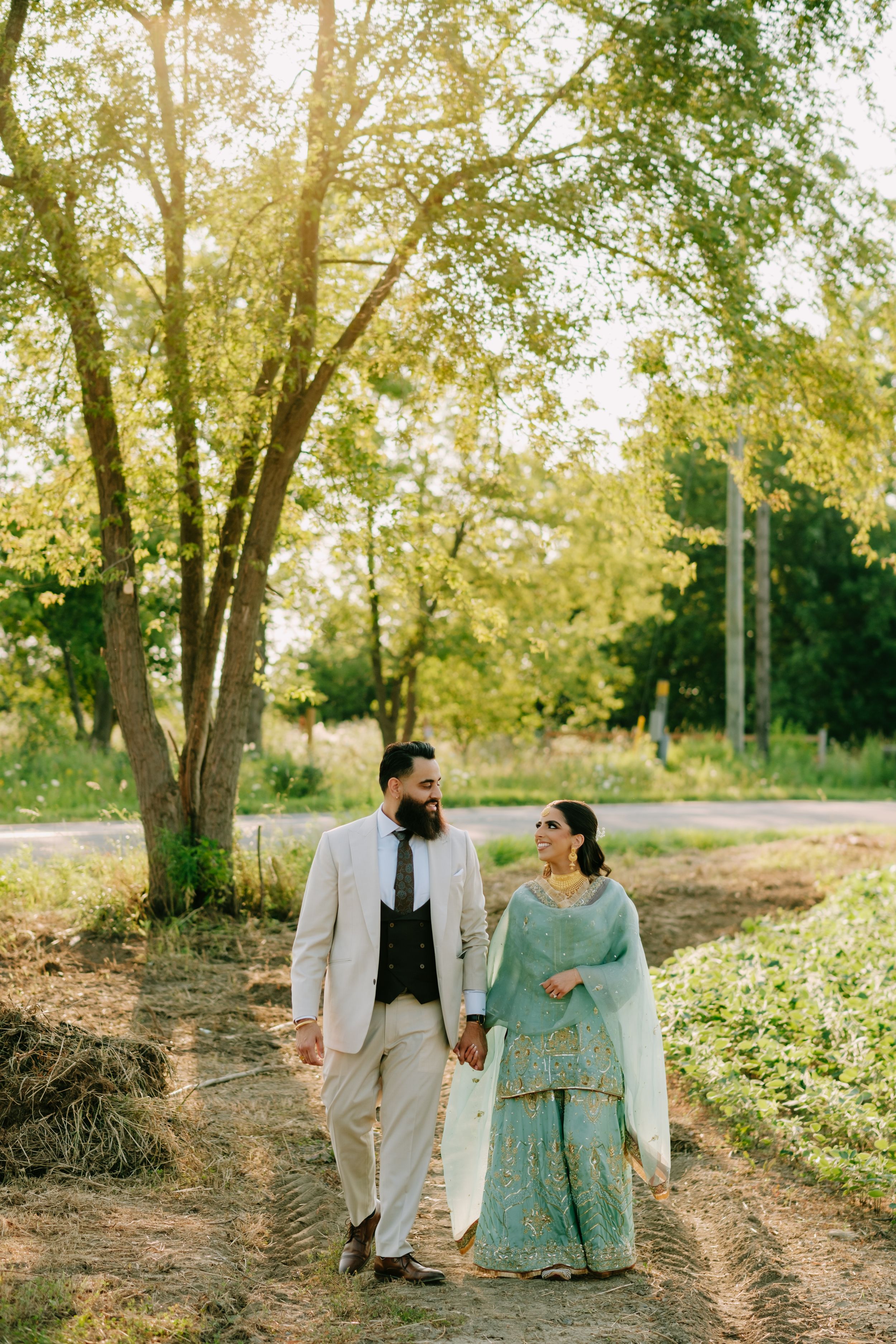 A couple walks hand in hand on a dirt path surrounded by sunlit trees, wearing elegant traditional South Asian attire.