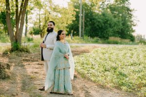 A couple in traditional South Asian wedding attire stand together on a sunlit path surrounded by trees and greenery.