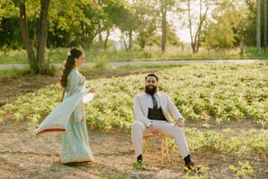 A couple in formal South Asian wedding attire pose playfully on a dirt path with trees in the background.