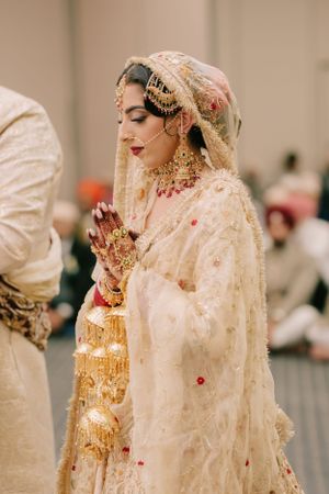 Traditional South Asian bride in cream and gold embroidered outfit with ornate jewelry and henna adorned hands during ceremony.