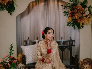 Traditional South Asian bride in ornate gold lehenga smiles while seated at decorated wedding venue with autumn flowers.