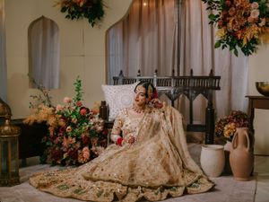 A person in an ornate cream-colored lehenga sits elegantly against pillows surrounded by floral decorations and vases.