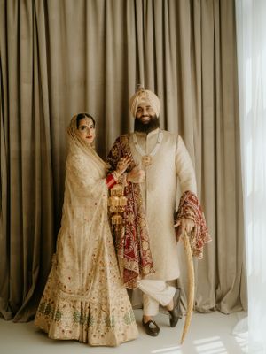 Traditional Indian wedding couple in cream and gold attire posing against draped curtains.
