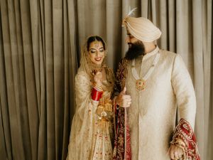 A Sikh couple in traditional cream and gold wedding attire share a joyful moment together during their ceremony.