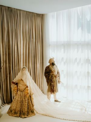 Traditional Sikh wedding outfits displayed in soft lighting with gold lengha and flowing veil against beige curtains.