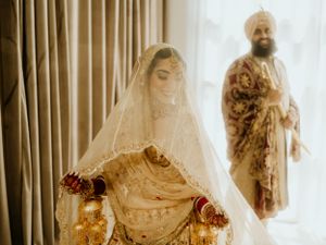 Traditional Indian wedding couple in elegant cream and gold outfits pose during ceremony with soft lighting and drapes.