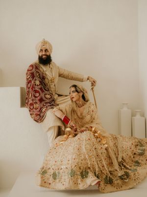 Traditional Sikh couple in ornate wedding attire featuring cream and gold lehenga and turban in minimalist setting.