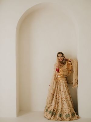 Bride in ornate cream and gold lehenga poses gracefully in minimalist archway setting.