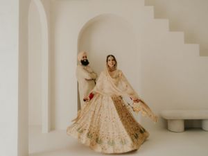 A person wearing a traditional cream and gold embellished lehenga twirls in an elegant pose against white architectural walls.