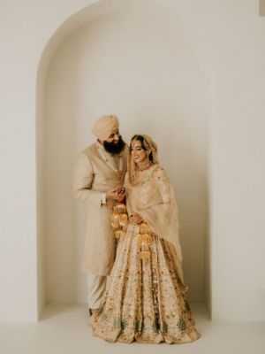 Elegant Sikh wedding couple in matching cream and gold attire pose together in front of arched backdrop.