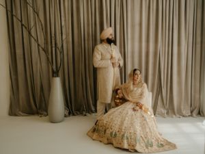 A couple in traditional Indian wedding attire poses elegantly against a neutral backdrop with draped curtains and a tall vase.