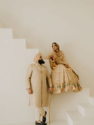 A couple in traditional wedding attire posed against a white wall with stairs in a minimalist setting.