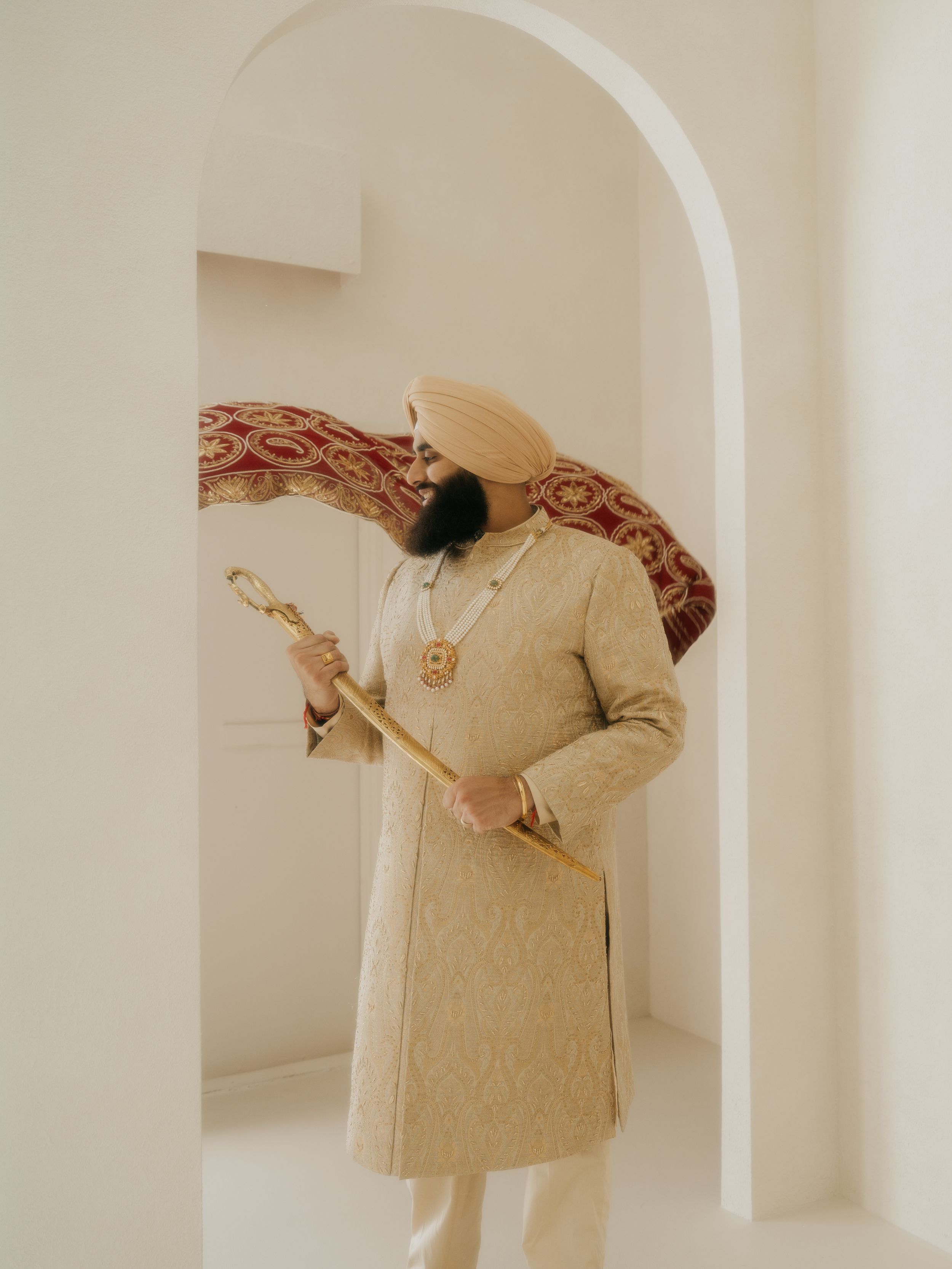 Traditional groom in cream sherwani holds ceremonial sword while standing in white arched doorway.