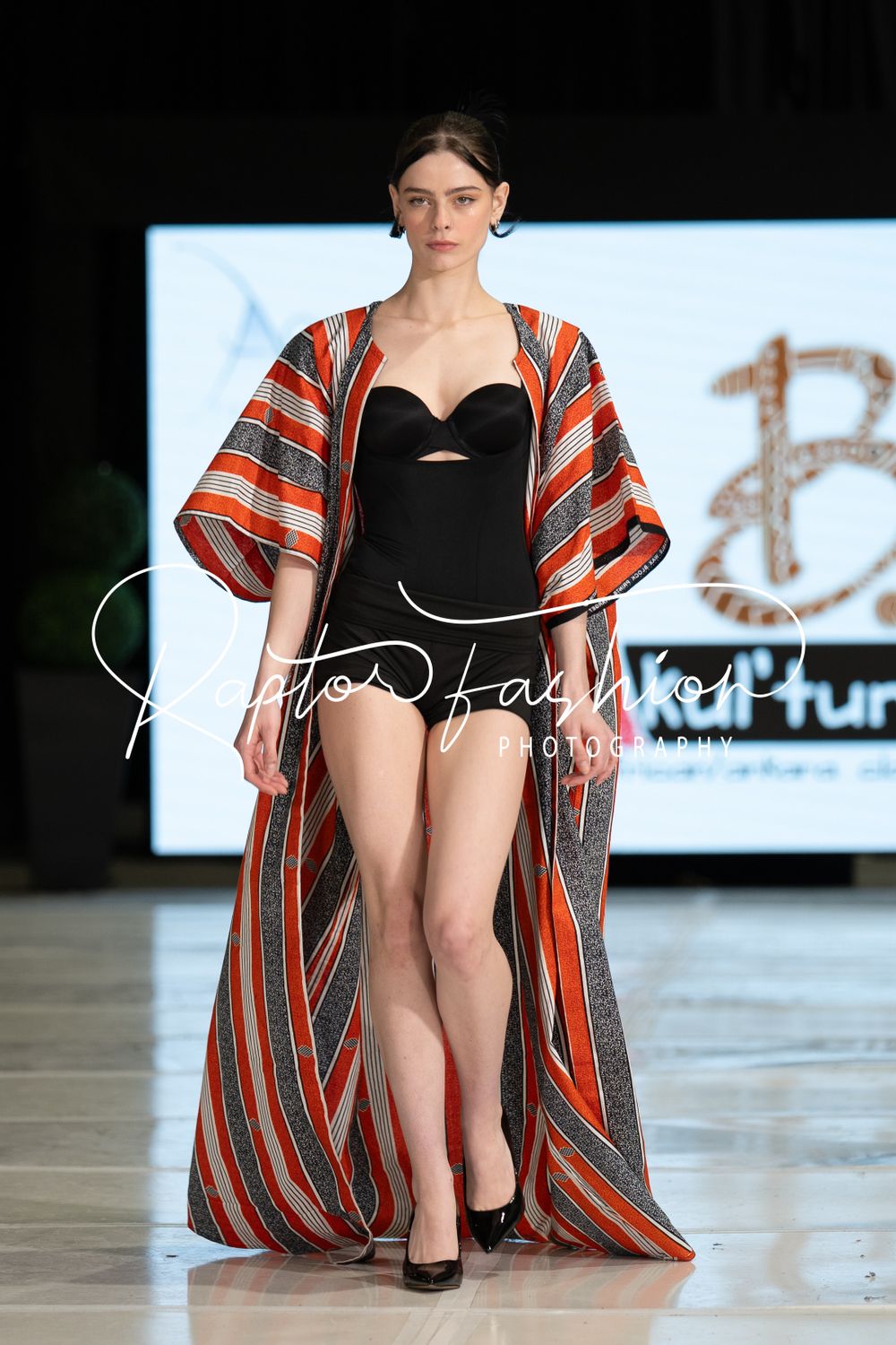 Model walks runway wearing black swimsuit with orange and grey striped dramatic floor-length kimono cover-up at fashion show.