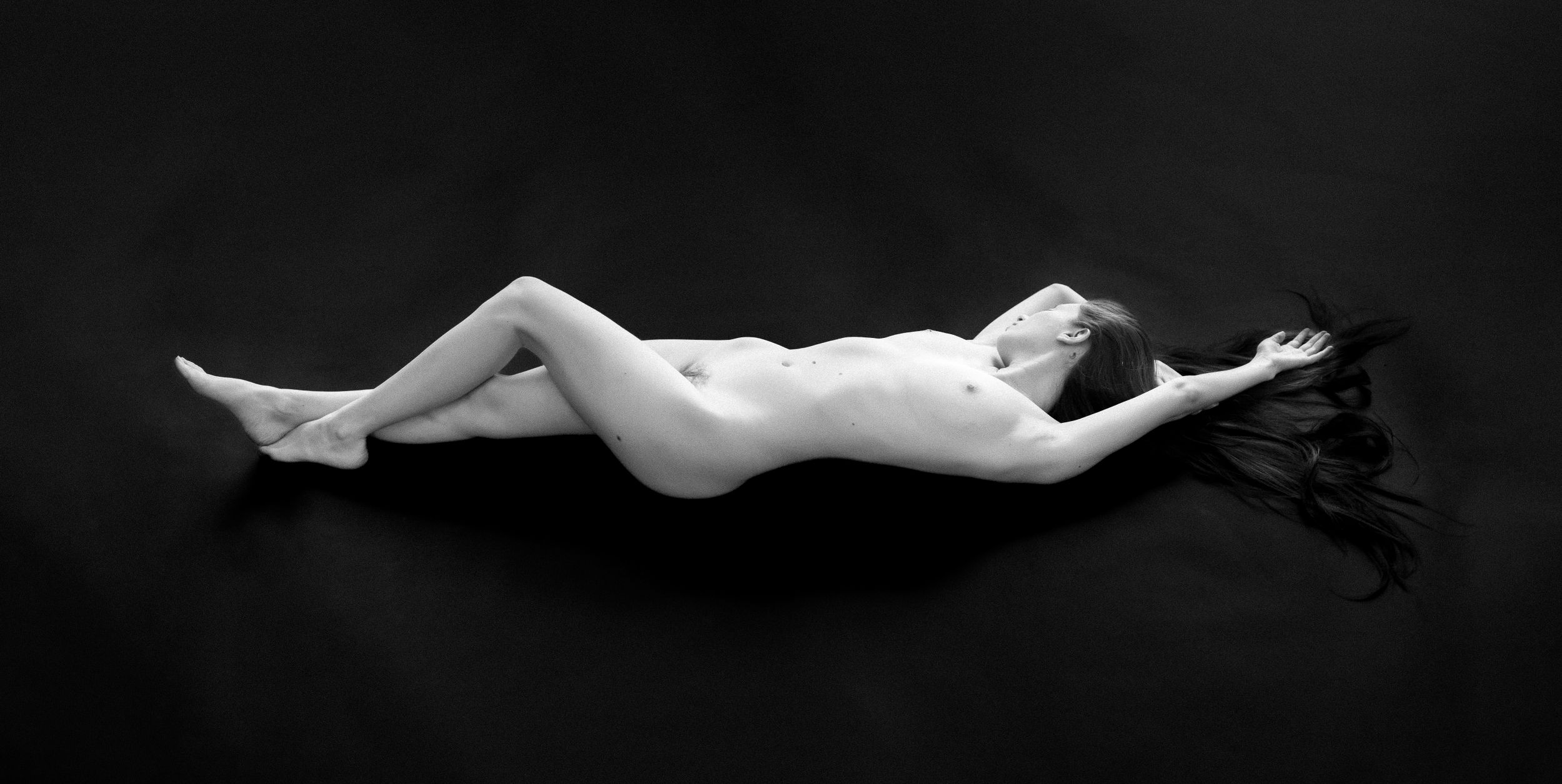 Black and white artistic nude study showing a reclining figure against dark backdrop.