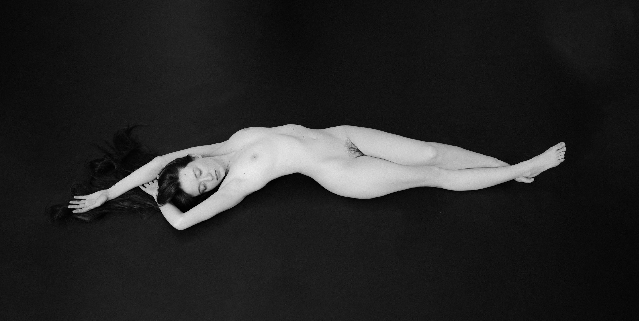 Monochromatic photograph of a nude figure in graceful reclined pose on dark background.