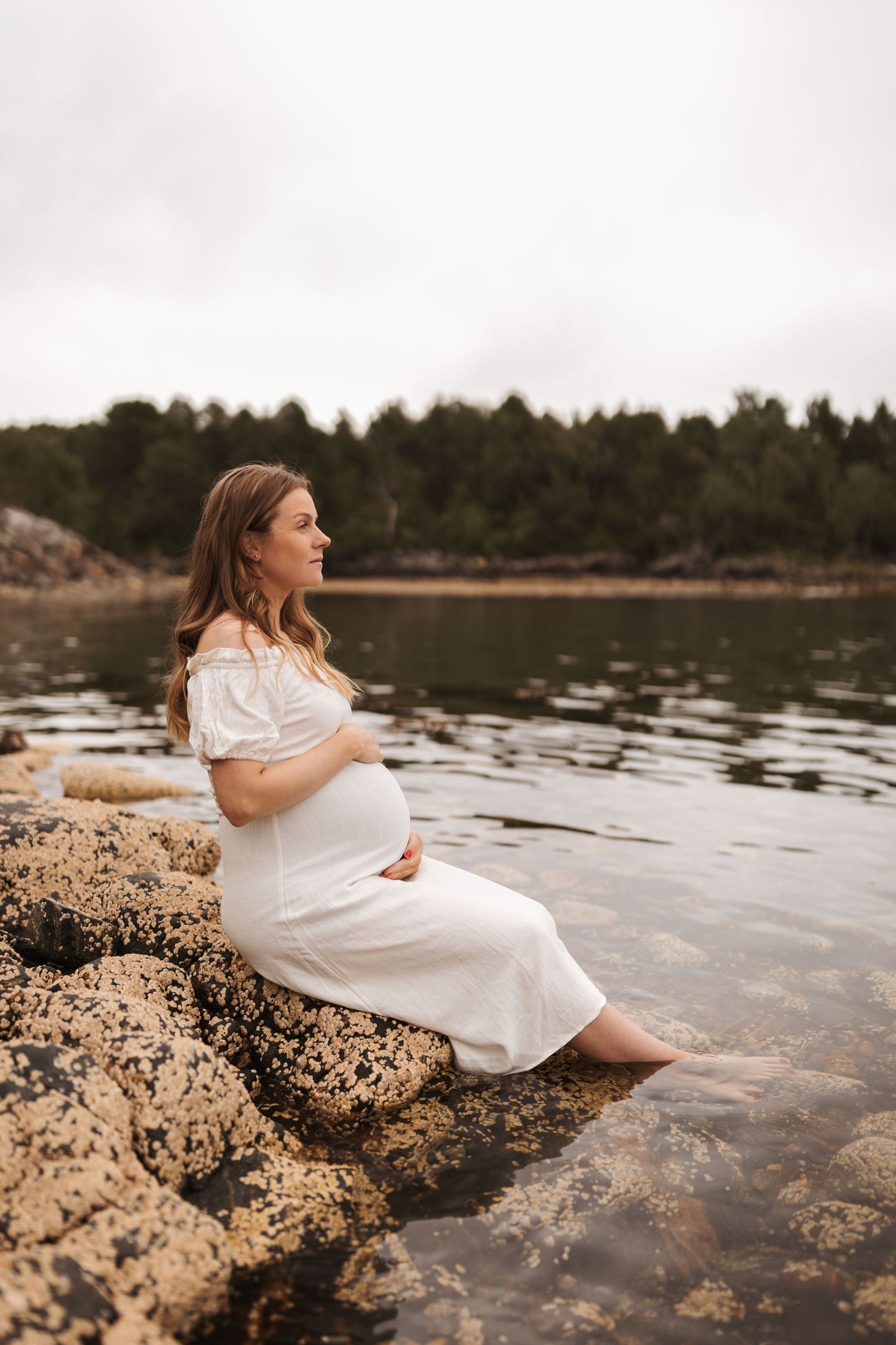 Pregnant woman in white dress sitting on rocky shore at sunset, dipping feet in calm lake waters.
