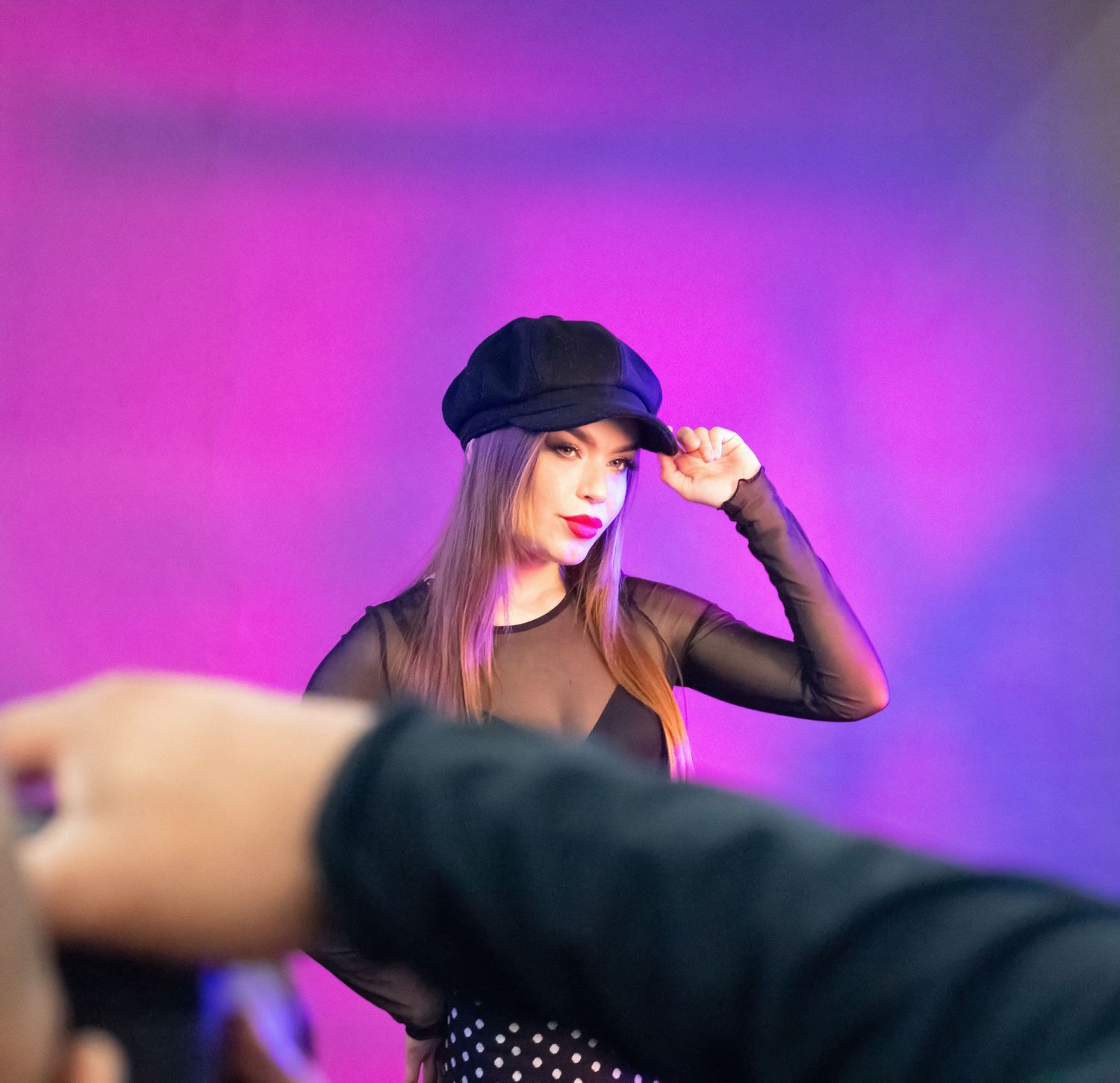 A fashion model in a dark outfit and hat poses against a vibrant purple backdrop during a photoshoot.