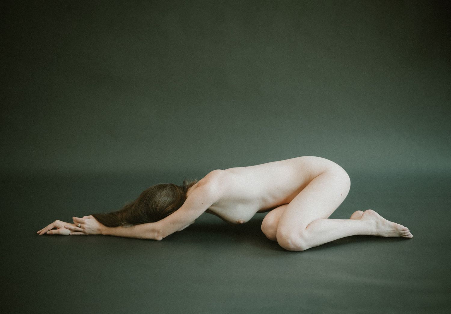 An artistic nude figure kneels in a graceful pose against a dark green backdrop in a minimalist studio setting.