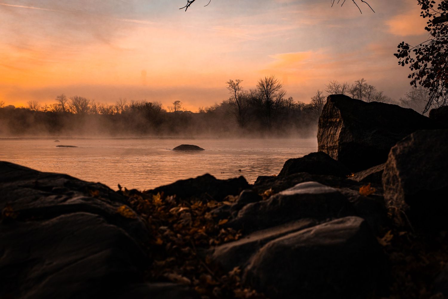 Misty lake shore with rocky outcrops silhouetted against an orange sunset sky and bare trees in the background.