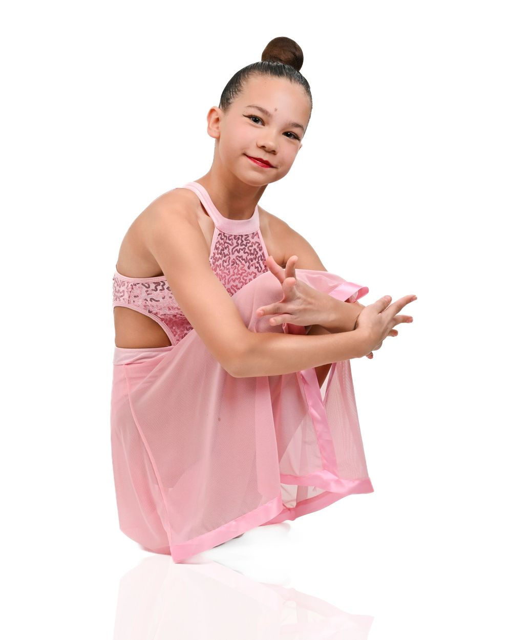 Young dancer in a pink sequined dance costume strikes a graceful seated pose against white background.