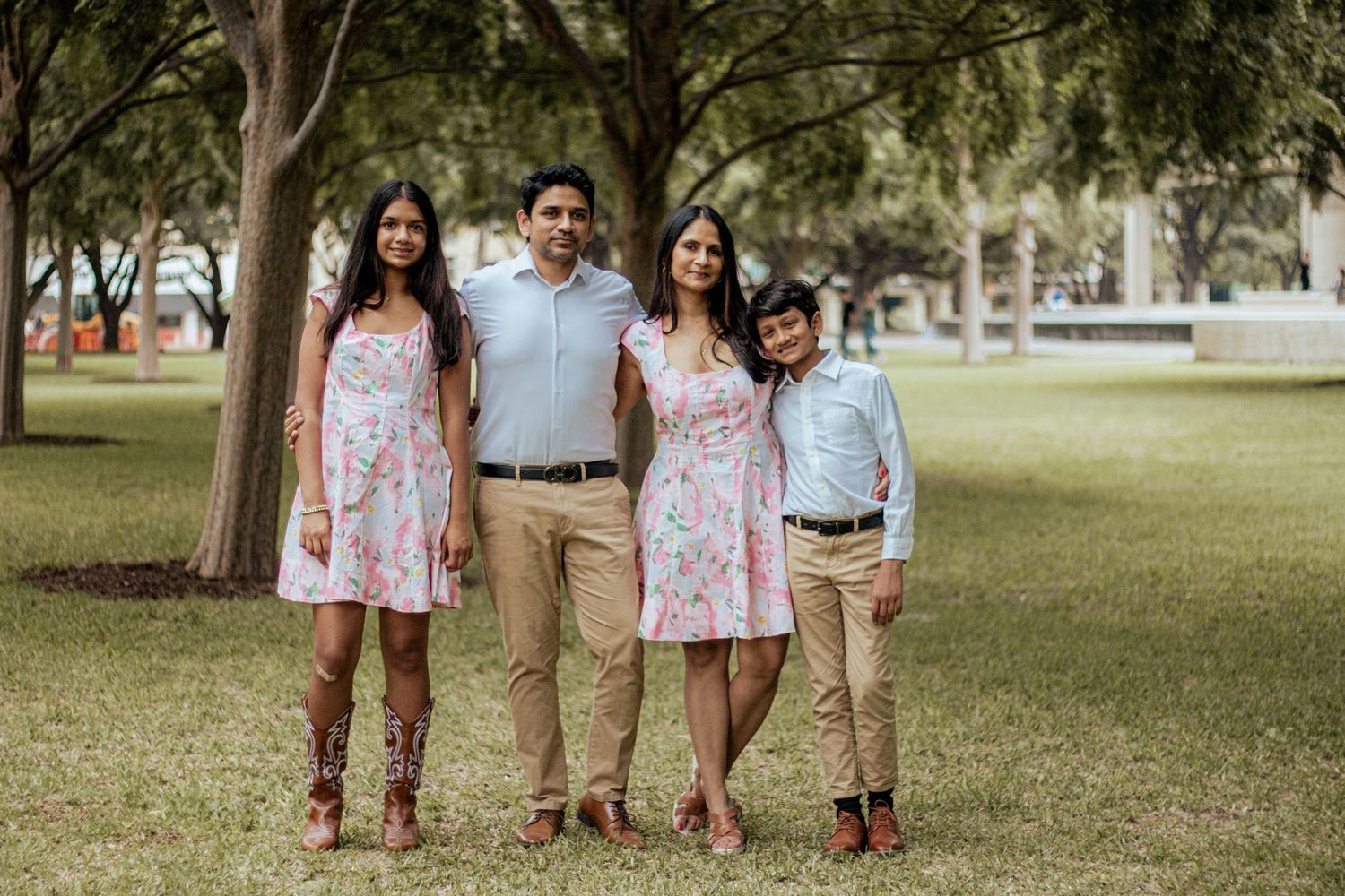 A family of four stands together on grass wearing coordinated pink and khaki outfits in a park setting.