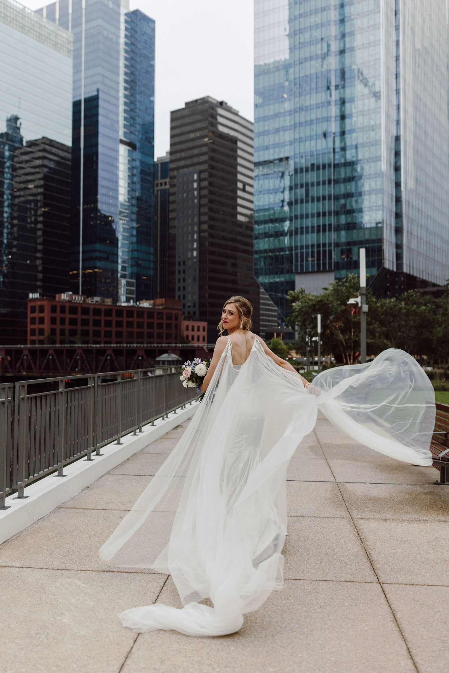 A flowing white wedding dress and veil billow in the wind against a backdrop of modern city buildings.